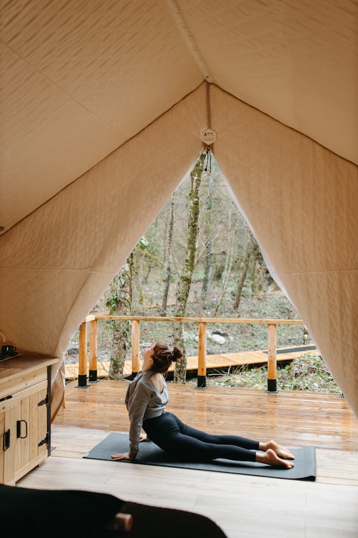 A woman stretches in a tent, practicing yoga on a mat in an outdoor forest setting.